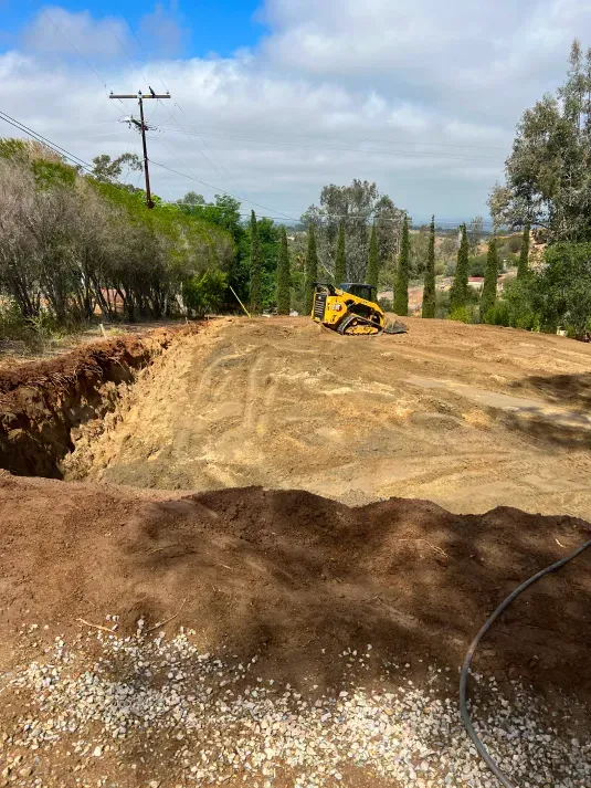 Yellow skid steer on a dirt worksite, cut-out pit in foreground, trees, and blue sky.