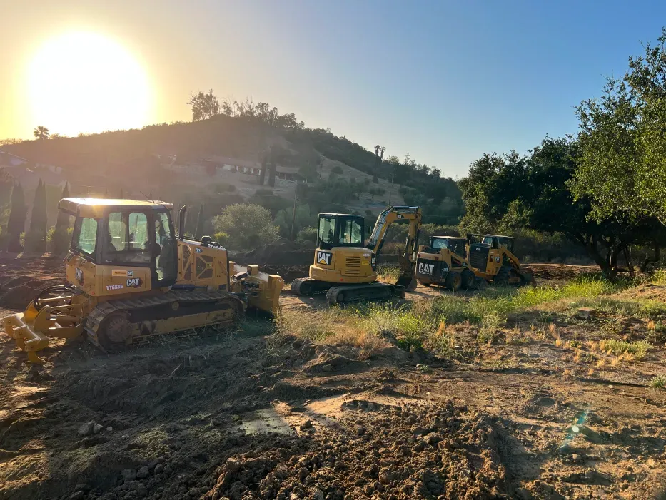Construction equipment parked in a field at sunrise, with a bulldozer and excavator visible.