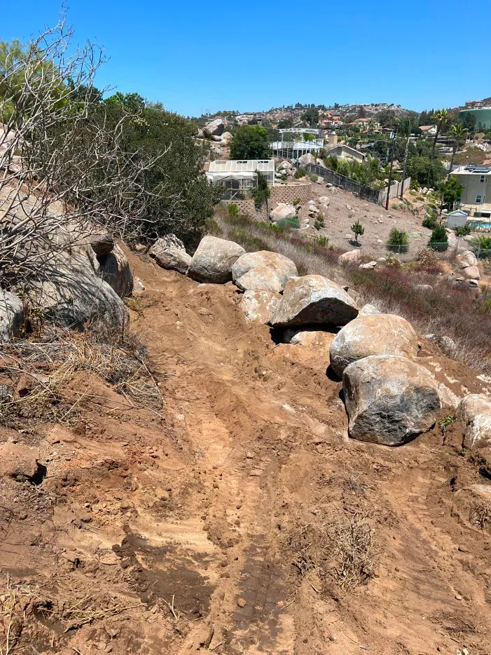 Dirt path with large rocks along a hillside, houses in the background under a clear blue sky.