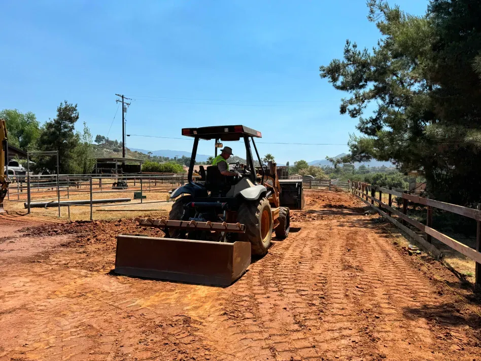 Tractor leveling a dirt road on a sunny day, a person driving. Fencing and trees border the road.