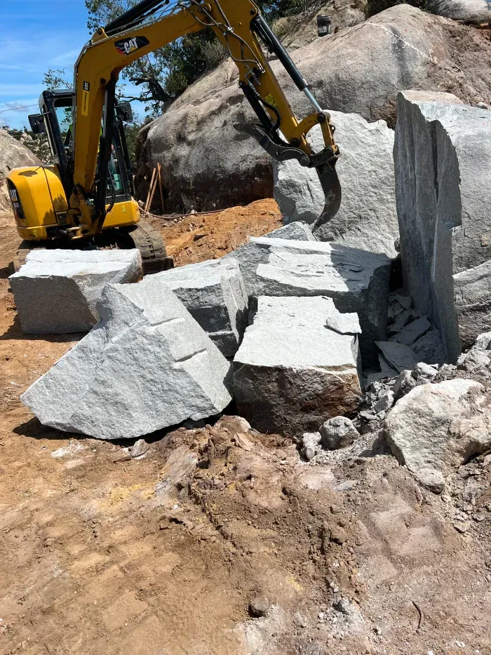 Yellow excavator breaking large gray rocks on a sunny day.