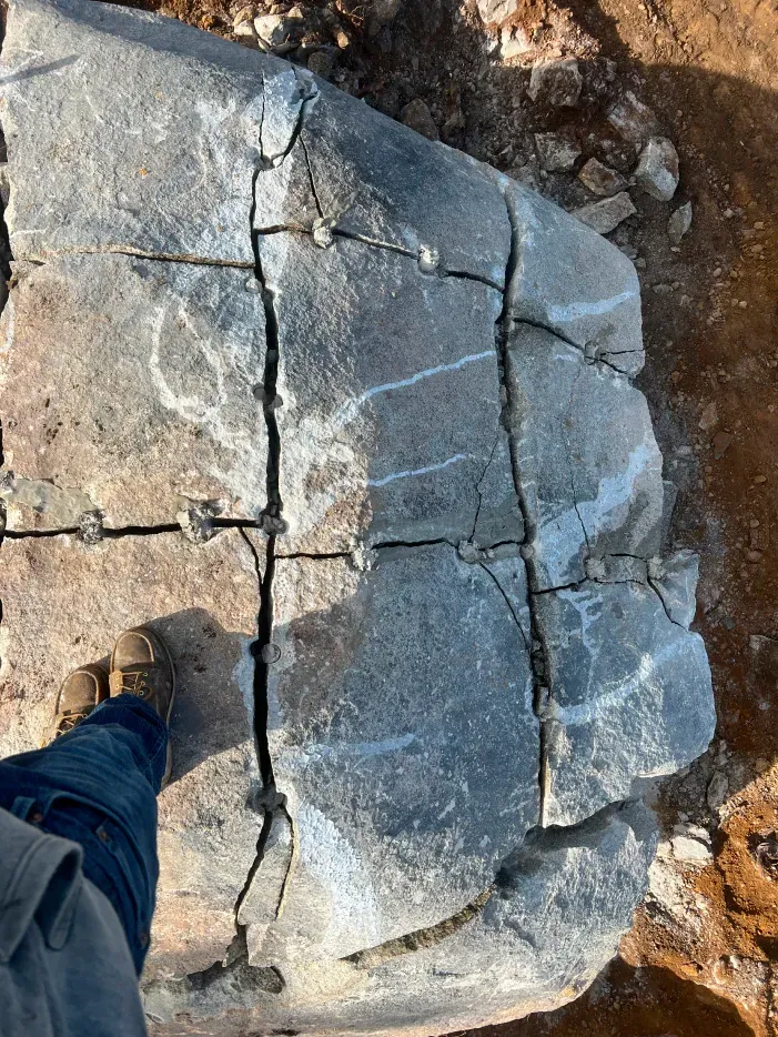 Large gray rock with visible cracks and metal fixtures, foot in jeans visible. Outdoors.