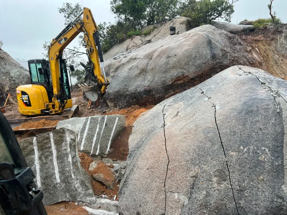 Yellow excavator breaking large granite boulders in an outdoor setting.