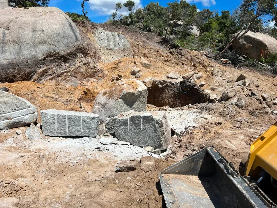Granite blocks cut with parallel lines on a hillside, next to dirt and an excavator.
