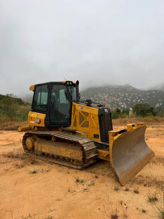 Yellow bulldozer on dirt, with a hillside and overcast sky in the background.