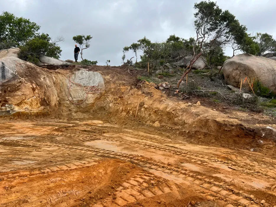 Dirt road with tire tracks leads to a hillside with rocks and a person standing near trees under a cloudy sky.