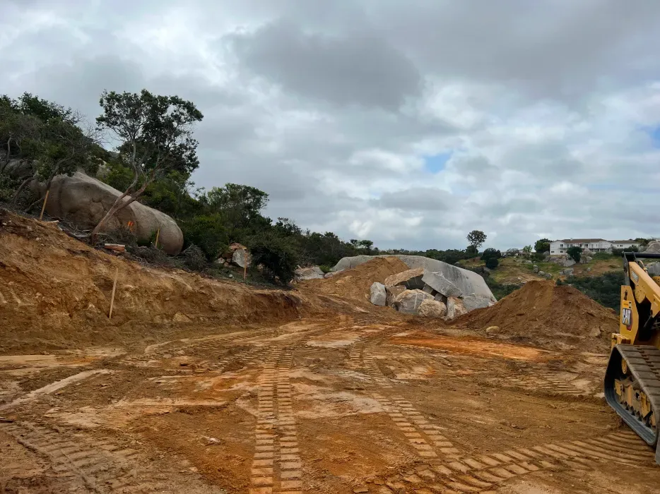 Construction site with dirt, heavy machinery, and large rocks under a cloudy sky.