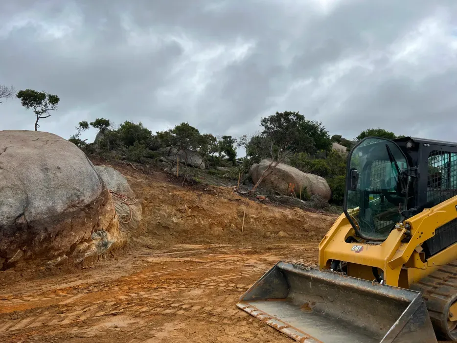 Yellow skid steer next to a rocky hillside under a cloudy sky, clearing land.
