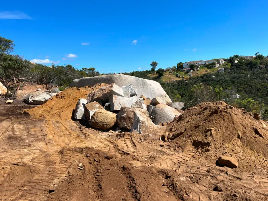 Construction site with dirt piles, rocks, and partially-cleared hillside under a blue sky.