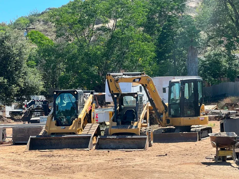 Three yellow construction vehicles parked on dirt: a skid steer, a mini-excavator, and another skid steer.