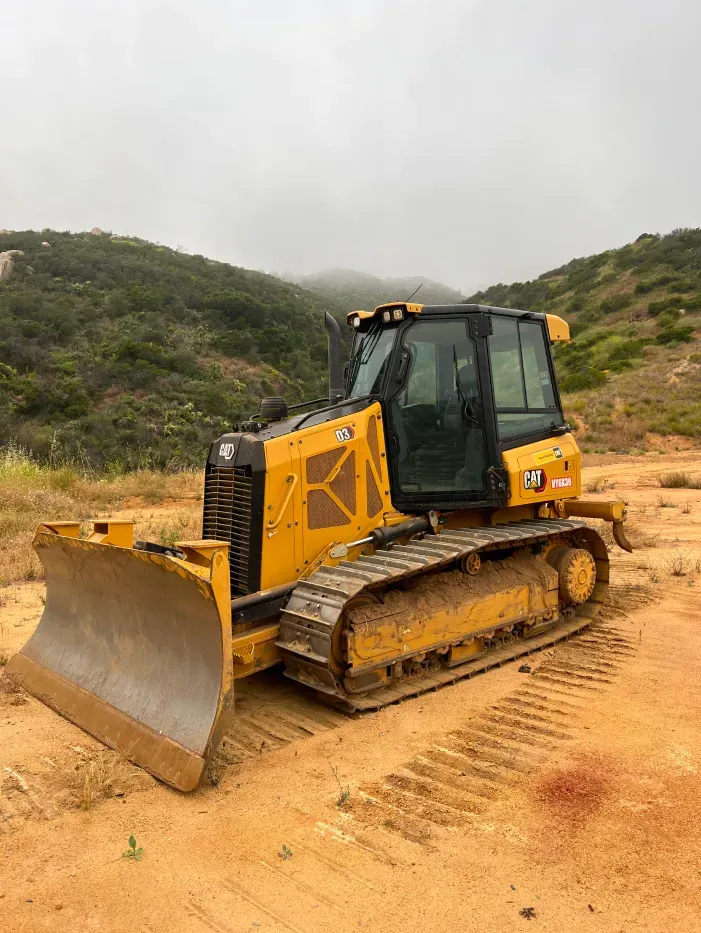 Yellow bulldozer on a dirt path, preparing to move soil with a backdrop of grassy hills under a cloudy sky.