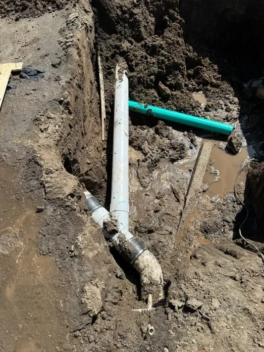 Pipes in a trench, with a broken section. Mud and dirt surround the pipes. Green pipe also in the trench.