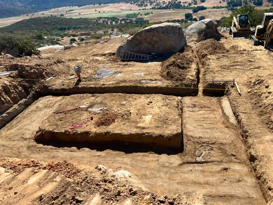 Archaeological dig site with rectangular trenches, heavy machinery, and a person.