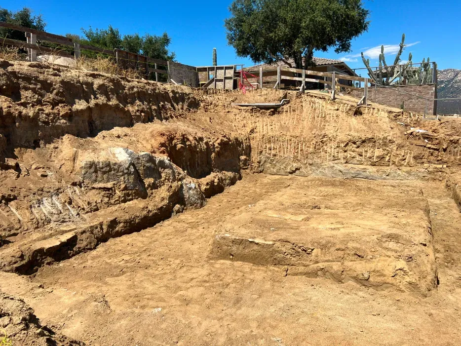 Excavation site with exposed earth, stone structures, and wooden fencing, under a clear sky.