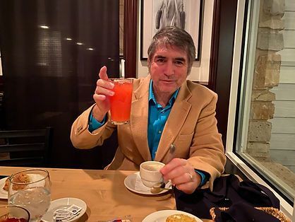 A person in a tan blazer and blue shirt sits at a restaurant table, holding a glass of orange juice and a small cup.