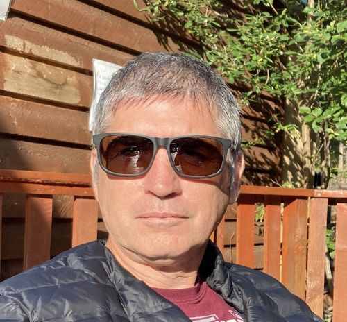A person with short grey hair wearing sunglasses and a dark puffer jacket sits outdoors in front of a wooden fence.