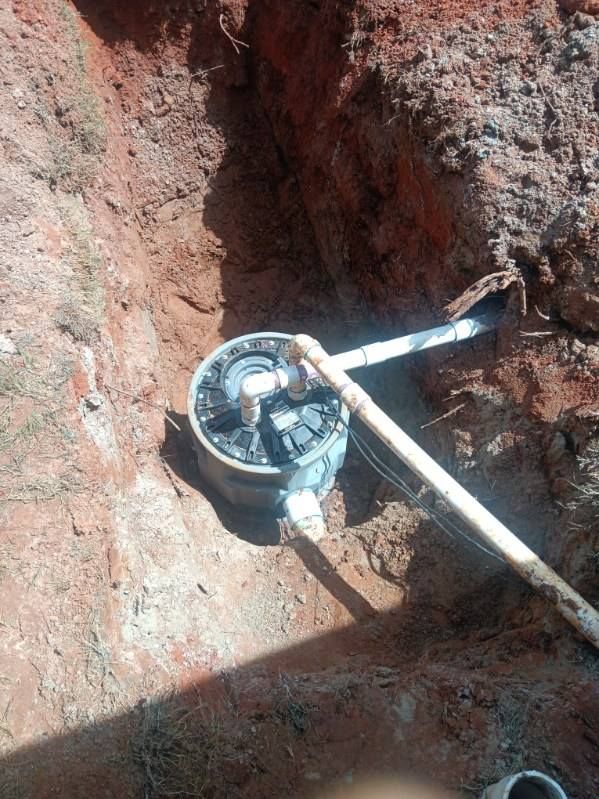 A circular septic system component is installed in a red-soil trench. PVC pipes extend from the component.