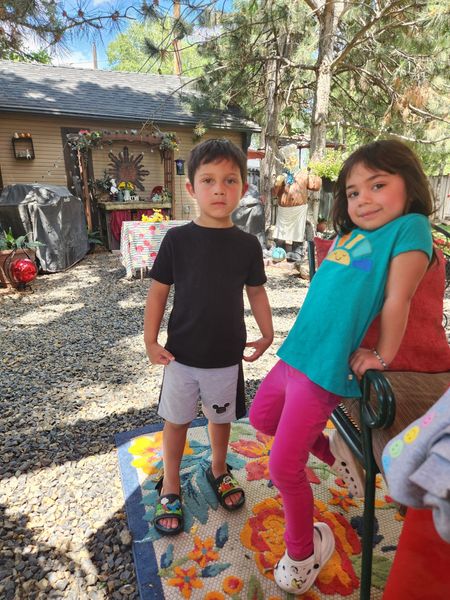 Two children, a boy and girl, pose outdoors. The girl wears pink leggings and smiles; the boy wears shorts and sandals.