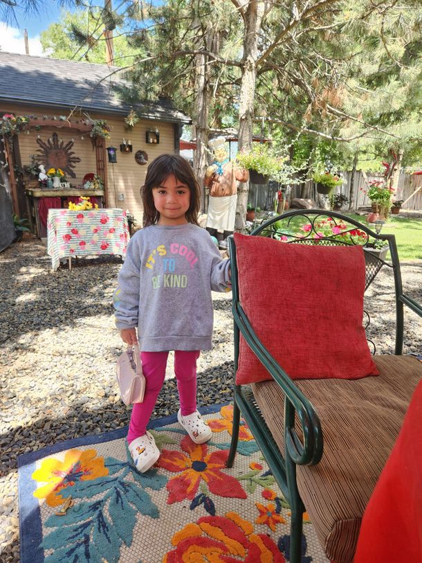 Girl in gray sweatshirt and pink leggings stands on a floral rug in a backyard.