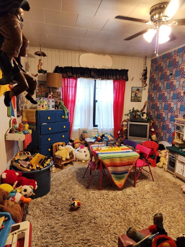 Cluttered children's room with toys, a window, and furniture. Shag carpet, red curtains, blue dresser.