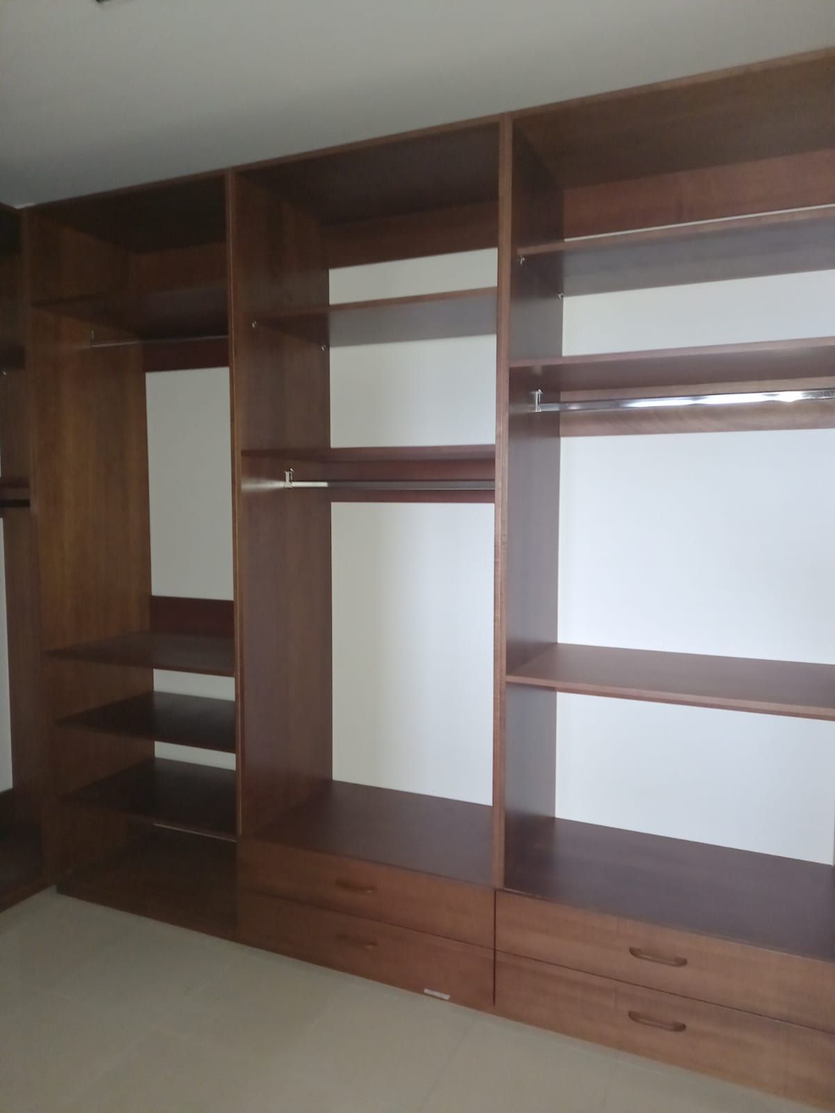 Armadio a muro in legno con ripiani aperti, cassetti e aste appendiabiti. Legno marrone contro pareti bianche.