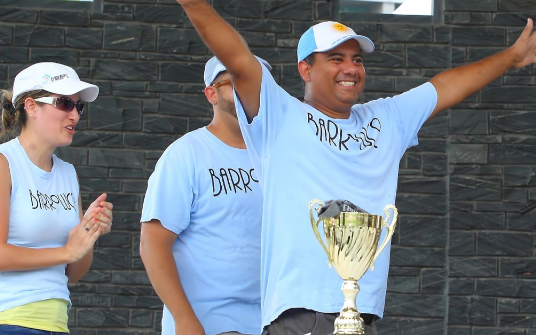 Man in light blue shirt raises arms, holding trophy, two people clap nearby.