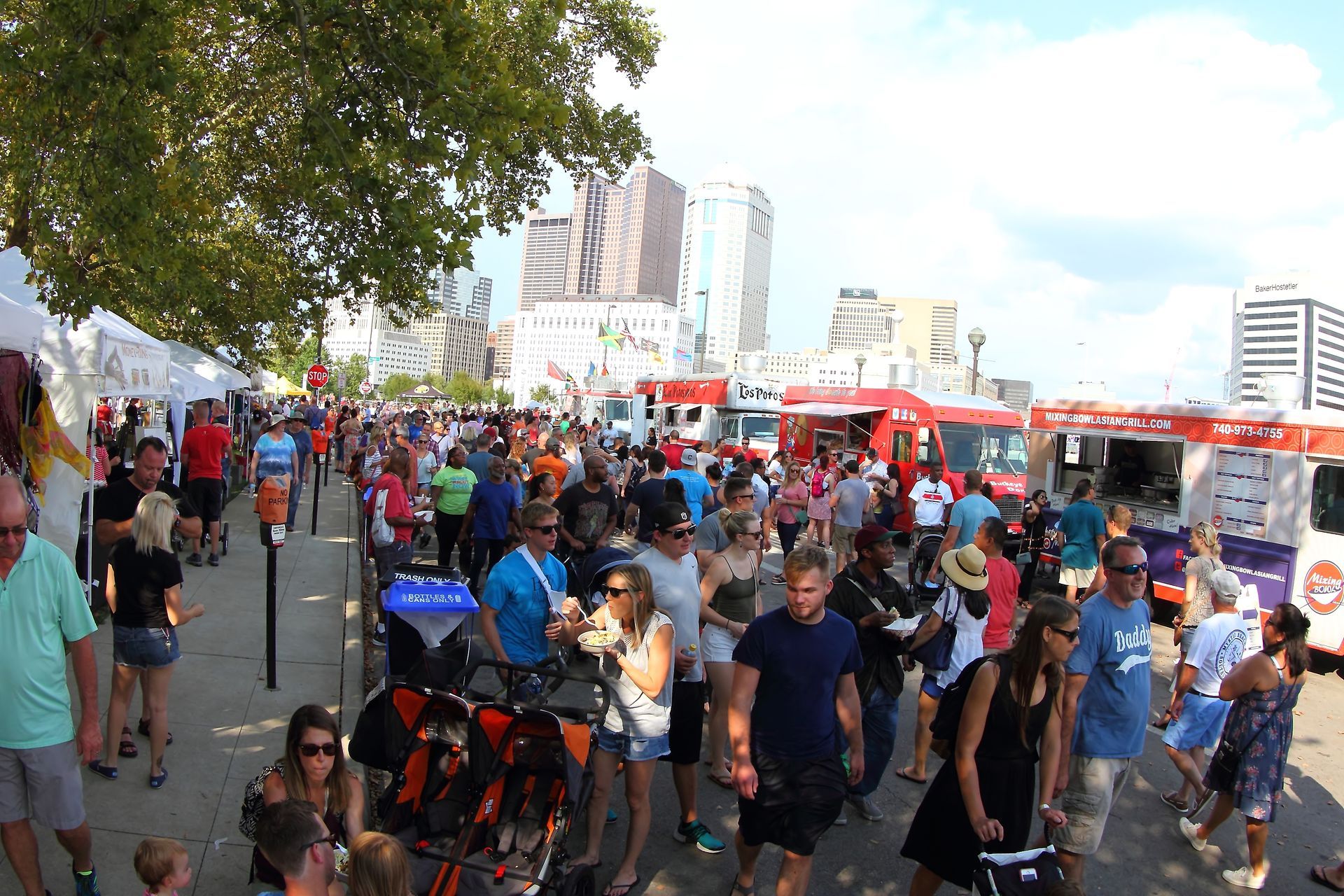 Crowd at a food truck festival with various vendors, in a city with tall buildings, sunny day.