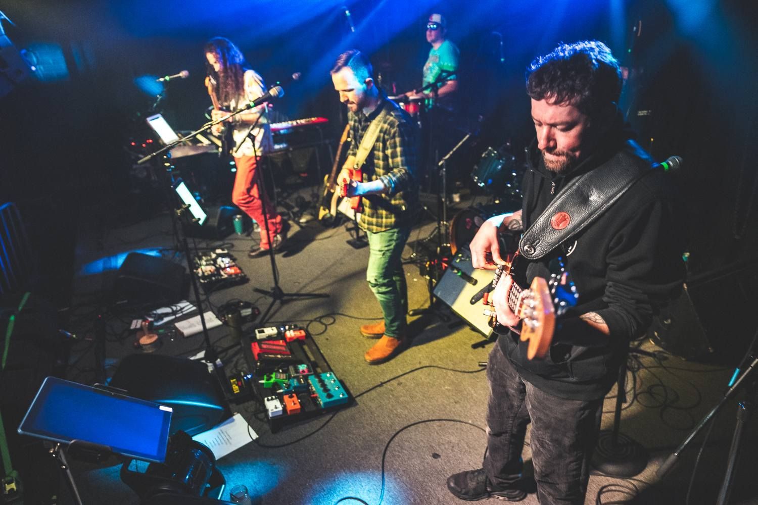 Band performing on a dark stage with blue lighting, guitars, microphones, and pedalboards visible.