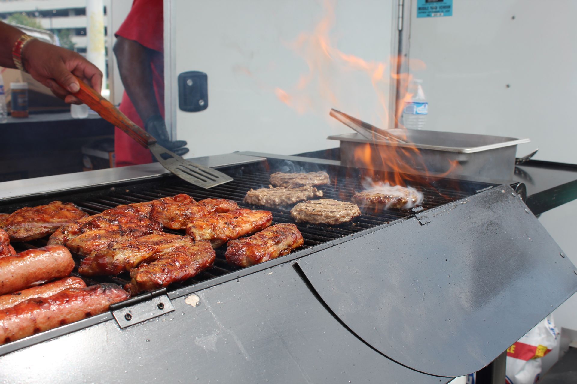 A person grilling food on a barbecue, including burgers, chicken, and sausages, with flames visible.