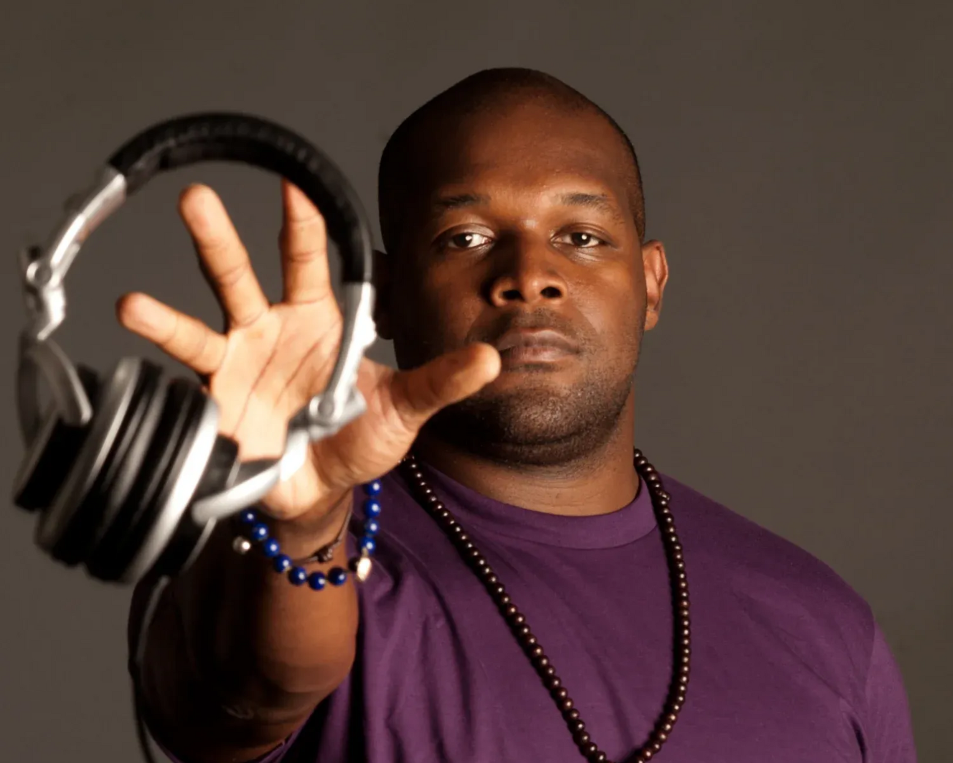 Man in purple shirt holding out headphones toward the camera against a dark background