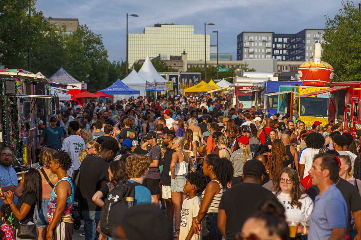 Crowded outdoor street festival with tents, food trucks, and people walking under a sunny sky