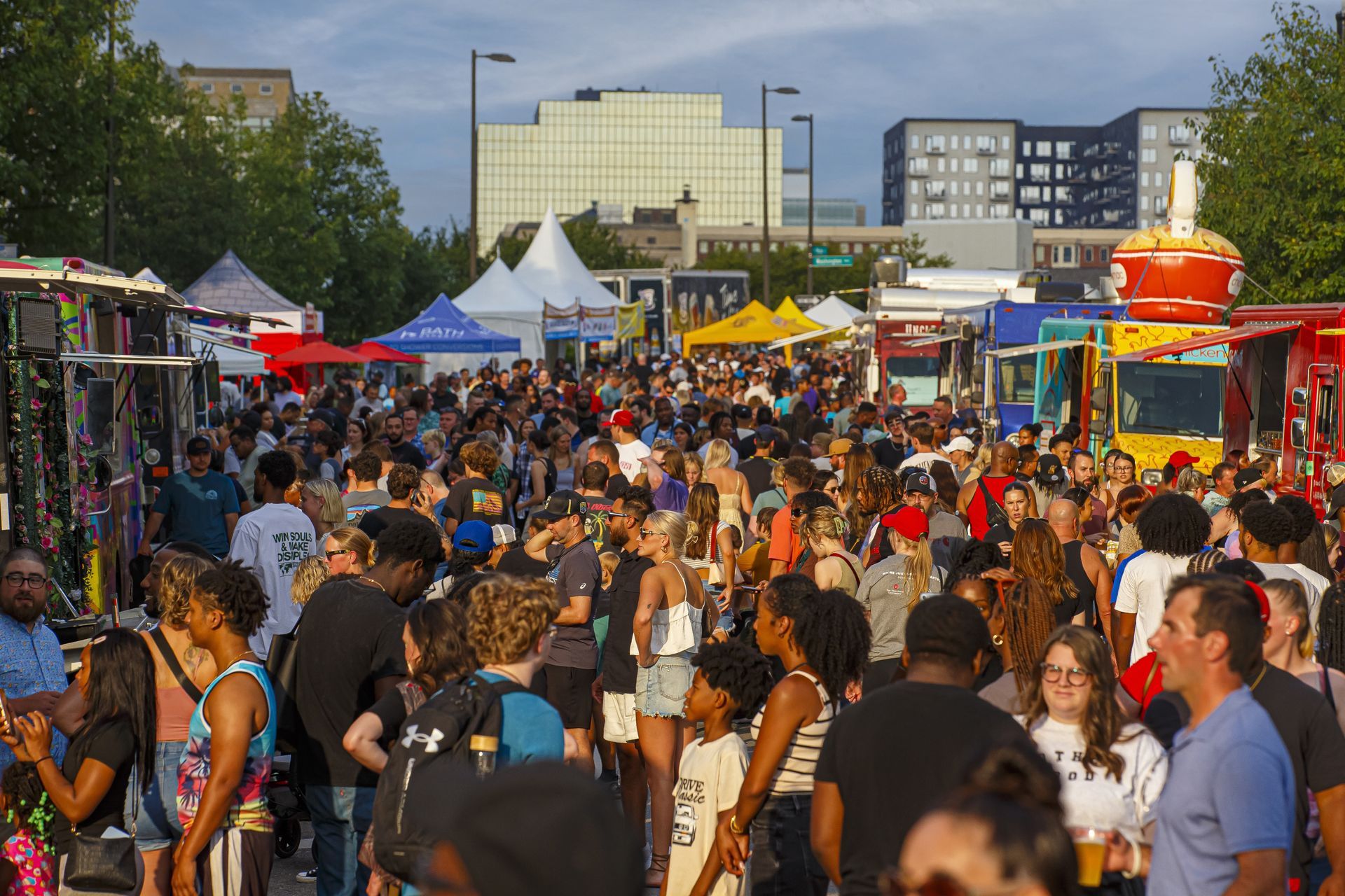 Crowd at a food truck festival with various trucks, tents, and buildings in the background.