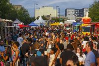 Crowd at a food truck festival with various trucks, tents, and buildings in the background.