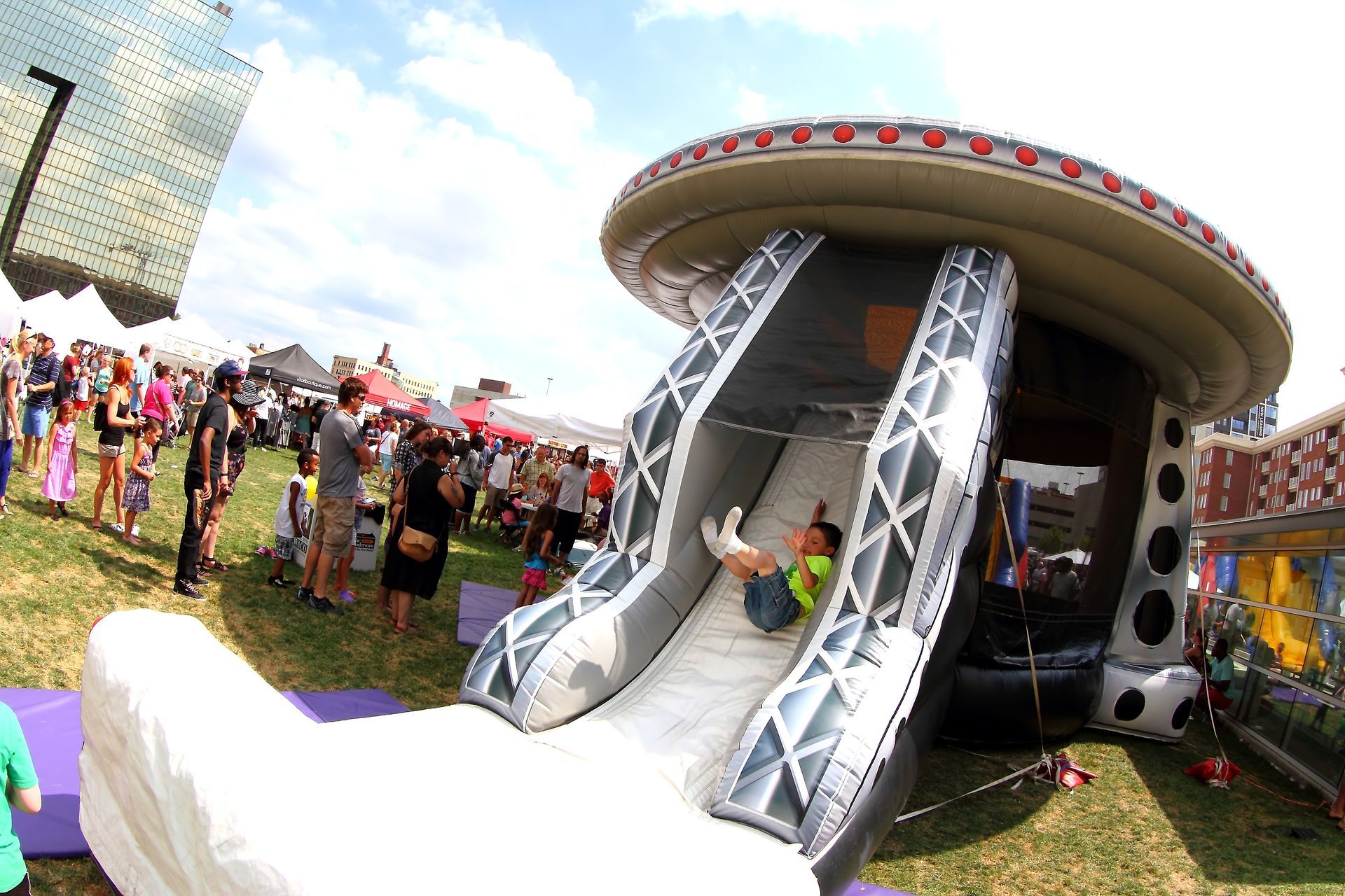 Child sliding down a black and white inflatable slide at an outdoor event, with a crowd of people in the background.