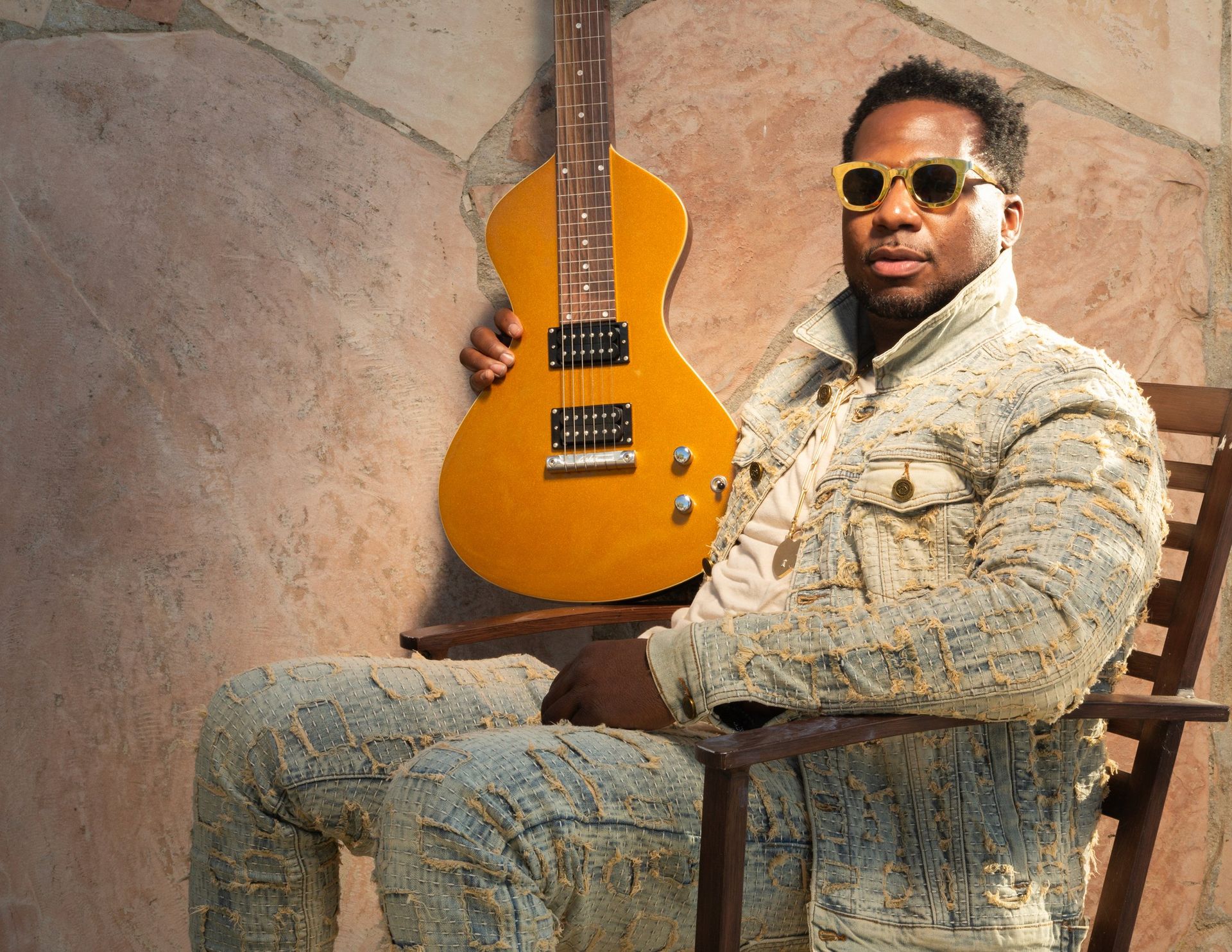 Man sitting in a chair, holding a yellow guitar against a textured wall, wearing sunglasses and a light suit.