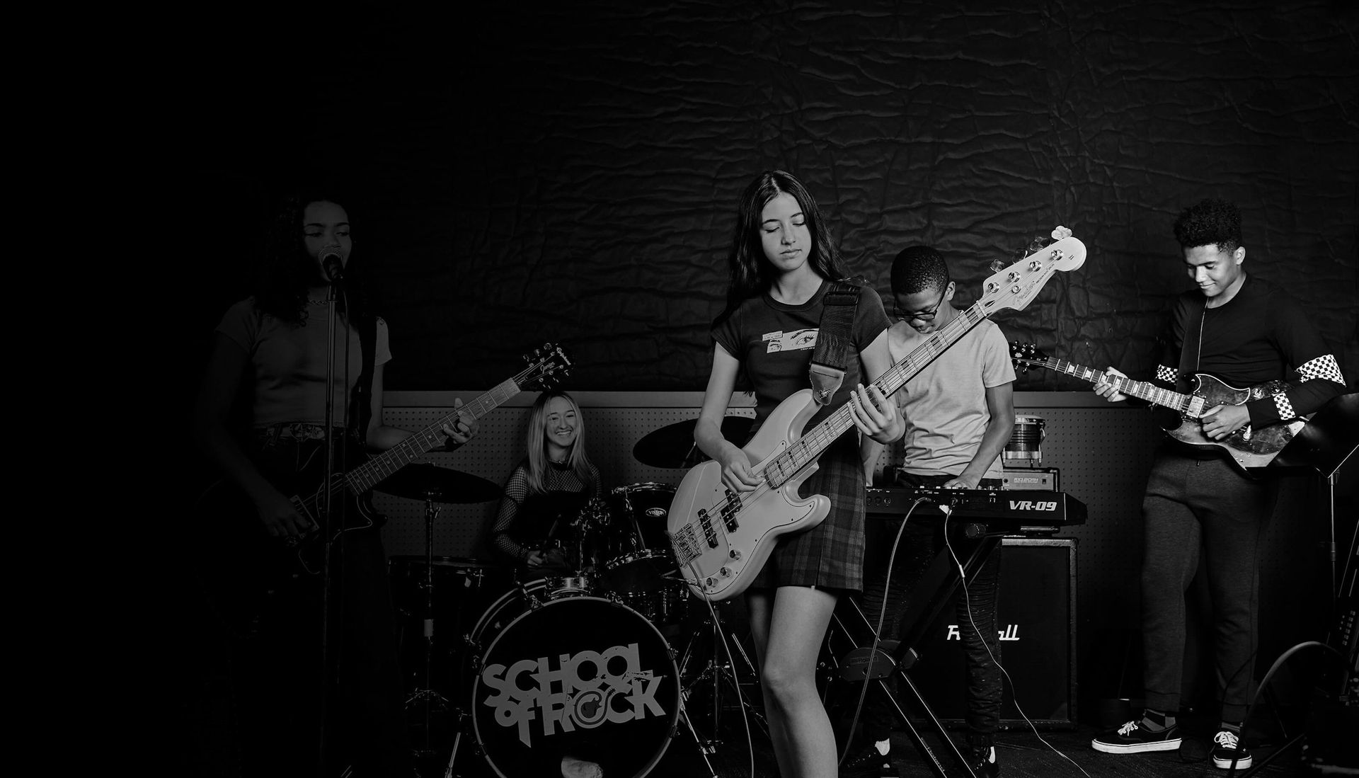 Band performing on a dark stage with blue lighting, guitars, microphones, and pedalboards visible.