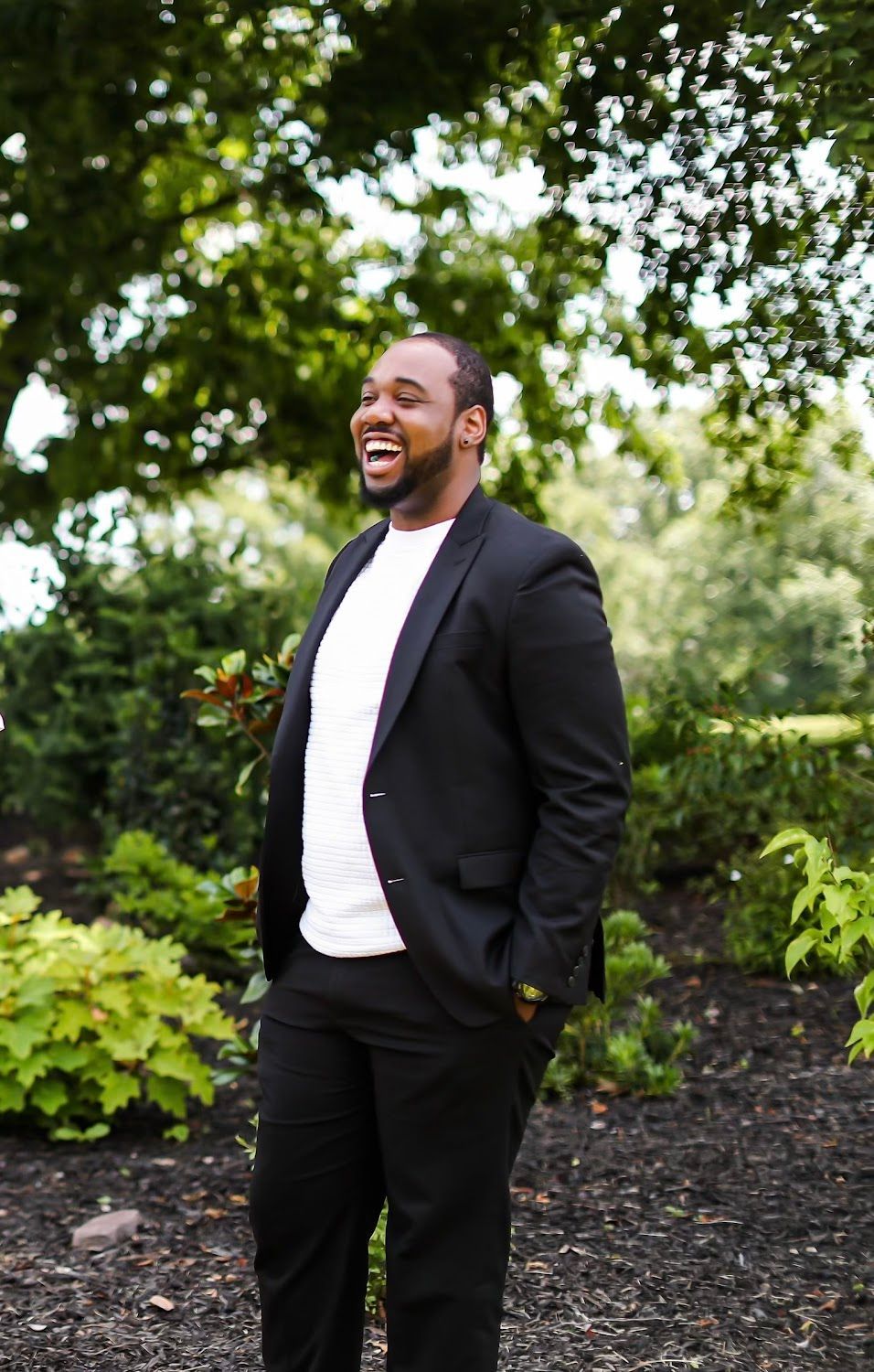 A man in a suit and white shirt is standing in front of a tree.