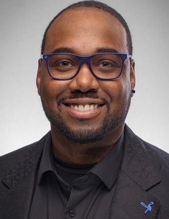 Man wearing blue-framed glasses smiles, dressed in a black shirt and blazer. Studio shot with gray background.