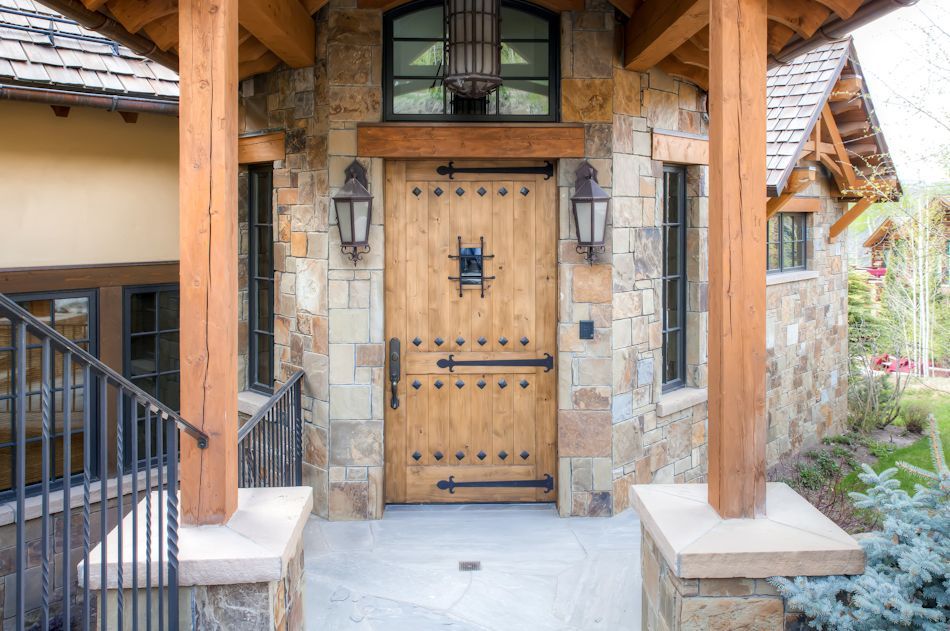 A rustic wood front door with black iron hardware in a stone entryway with wooden porch columns and a stone floor.
