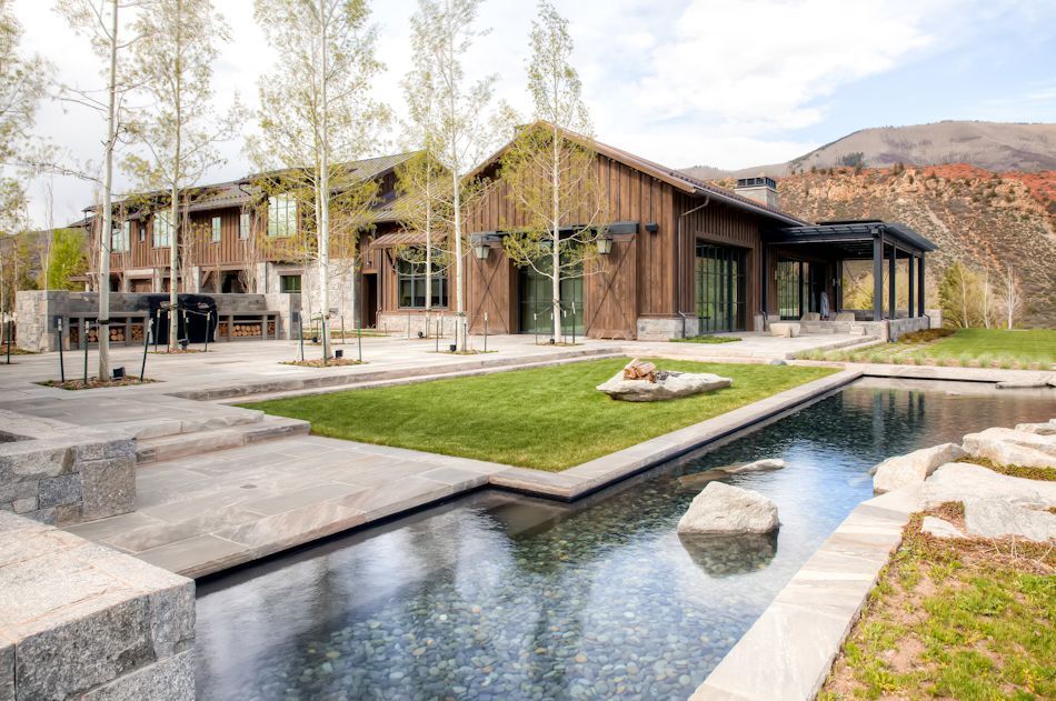 A modern rustic house with wood siding, a green lawn, and a reflective pool in a mountainous, sunny landscape.