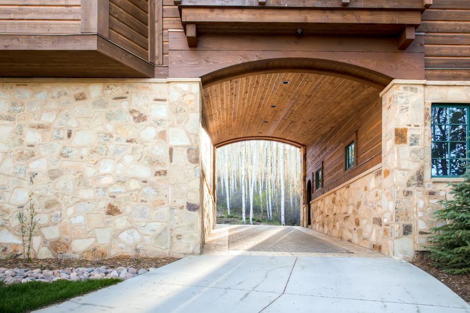 A stone and wood house with a large archway leading to a paved driveway and a forest of tall, white trees.
