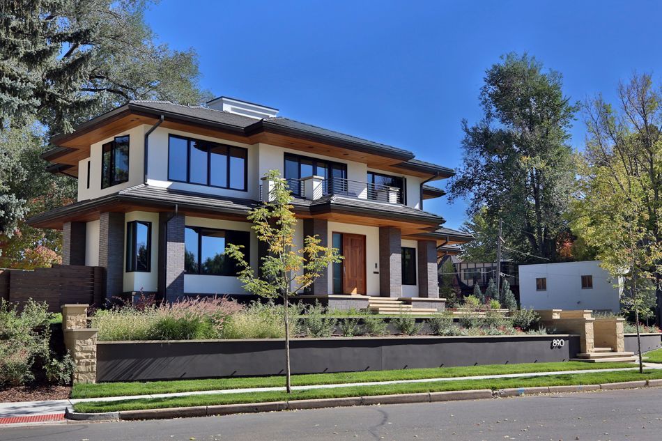 A two-story house with cream-colored stucco, dark trim, and a low-pitched roof, set behind a retaining wall and lawn.