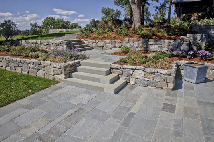 A stone-walled garden with purple salvia, red foliage, and pink flowers beside a stone staircase in a sunny landscape.