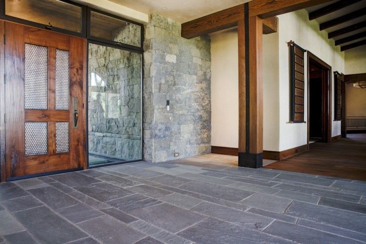 A bright, modern open-plan living room with grey sofas, a patterned rug, wooden coffee tables, and several green plants.