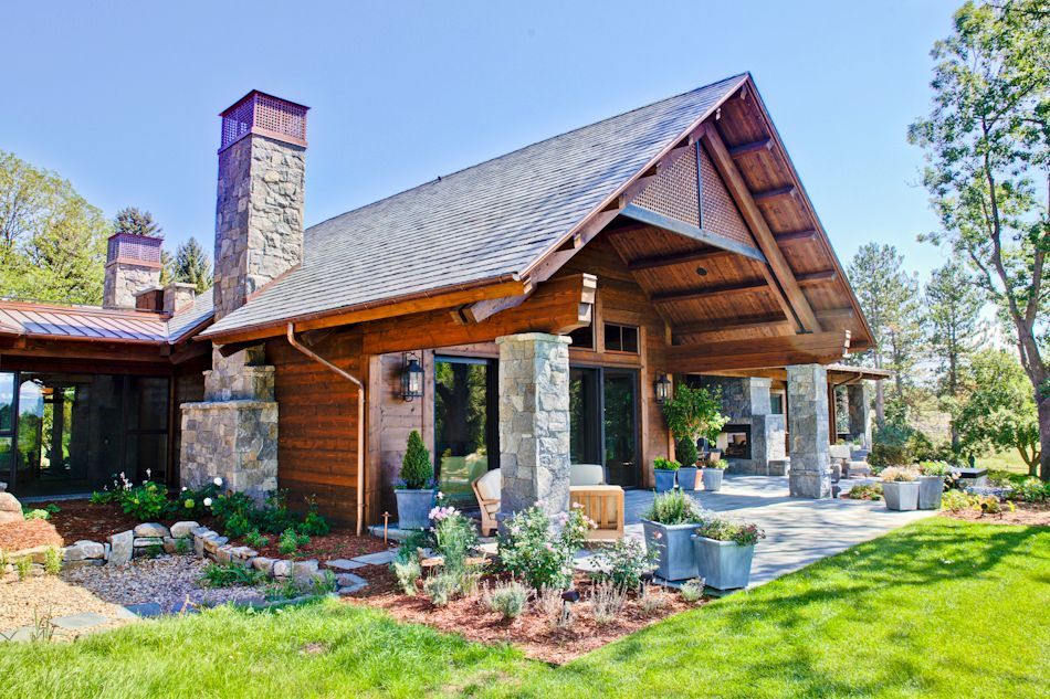 A rustic-style house with a covered patio, stone pillars, and a wooden exterior under a clear blue sky.