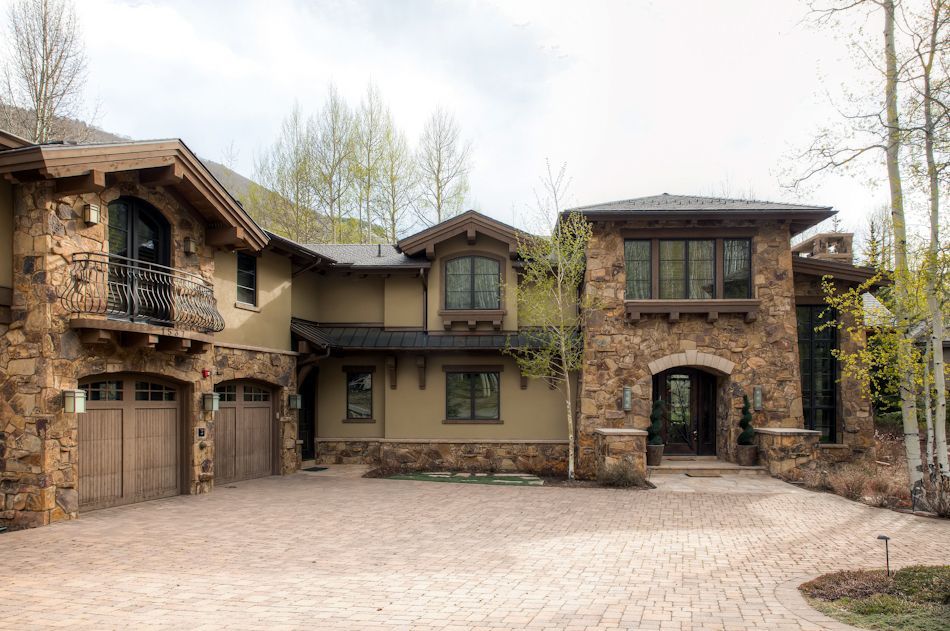 A large, two-story mountain home with stone siding, a dark roof, wooden garage doors, and a paver driveway.