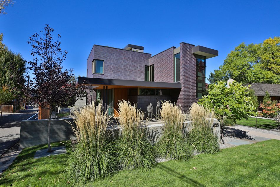 A modern, two-story house with dark, textured siding, wood accents, and large windows, surrounded by grass and tall grasses.