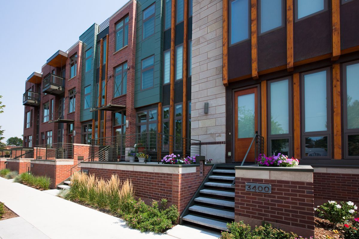 A multi-story townhome complex with red brick, glass panels, and stone accents, featuring outdoor staircases and planters.