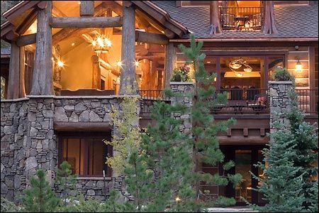 A multi-story rustic log cabin with stone masonry, large glass windows, and surrounding pine trees.