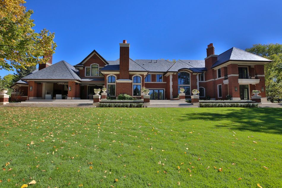 A large, multi-story brick mansion with a slate roof, situated behind a wide green lawn on a sunny day.